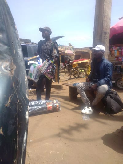The image captures an outdoor market scene in Mista Ali, Nigeria, under a bright, clear sky, with an uneven, reddish-brown dirt ground. In the mid-ground, a male subject stands, wearing a dark striped tunic and a dark baseball cap, holding multiple clear-plastic-wrapped bundles of small retail items. Adjacent to him on the ground are two large, dark, cylindrical packages, also plastic-wrapped with orange/red labels. To the right, another male subject sits on a rustic wooden bench; he wears a blue jacket, light jeans, white sneakers, and a white baseball cap. A black backpack rests on the ground near his right foot, and small blister packs are visible on the dirt in front of him. The background reveals a dark SUV parked behind the standing individual, a large concrete pillar with faded white markings, and various market stalls including one with a red canopy displaying numerous packaged goods. Several handcarts, some covered with yellow/orange tarps, are also present, along with simple, low-rise buildings with light-colored walls and some red roofs. A dark, weathered, curved object partially obstructs the left foreground.