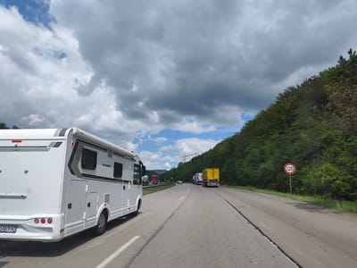 This is a point-of-view, landscape-oriented, medium shot of a highway in Gorno Kamartsi, Bulgaria. The scene is dominated by a white recreational vehicle (RV) on the left side of the frame. The RV has a license plate reading "5469 HO". In the background, the highway curves to the right, with several trucks and cars visible. The right side of the road is bordered by a dense forest on a hillside. A circular speed limit sign with the number "120" is mounted on a pole to the right of the road. The sky is cloudy with patches of blue. Power lines and pylons are visible in the distance.