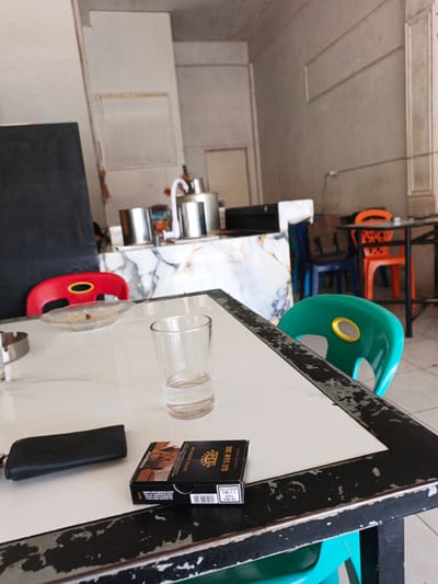 The image depicts a table in what appears to be a cafe or food stall in Geudonggeudong, Indonesia. On the table, there is a pack of "DJI SAM SOE" cigarettes, a black wallet or cardholder, and a glass of water. In the background, there are several colorful plastic chairs, including red, blue, green, and orange ones, arranged around tables. Some metal containers and a faucet are visible on a counter in the background, suggesting a preparation area. The overall environment appears to be functional and informal, consistent with a local eatery.