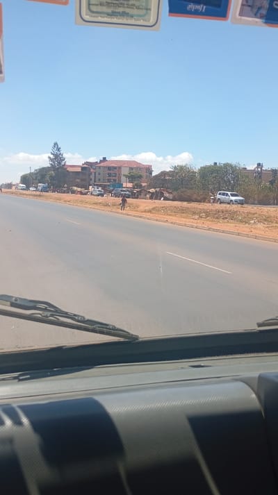 A sunlit, long asphalt road stretches into the distance. A man walks along the roadside, near buildings, vegetation, parked vehicles, and roadside stalls. The view is from the interior of a vehicle, with the dashboard and windshield wipers visible. The sky is clear and blue.