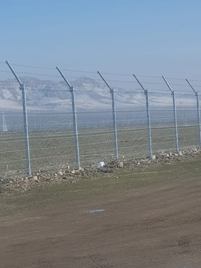 The image depicts an outdoor scene in Egizbulak, Uzbekistan. In the foreground, an unpaved dirt track exhibits tire impressions and a small water puddle. A strip of green grass and scattered dry leaves, along with a white plastic object, separate the track from a prominent metal security fence. The fence comprises multiple sections of welded wire mesh panels, supported by vertical posts, each featuring angled extensions at the top fitted with several strands of barbed wire. Through the fence, a utility structure, possibly a pylon base, is faintly visible on the left side. The background consists of distant mountains, partially covered in snow, set against a clear, pale blue sky with a slight haze near the horizon. No individuals are present, and no discernable actions or interactions are occurring.