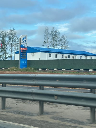The image captures a view of a Gazprom Neft gas station in the city of Bratovshchina, Russia. In the foreground, a metal guardrail is visible, suggesting the photo was taken from a moving vehicle. Behind the guardrail, a green hedge separates the road from the gas station premises.
The gas station itself features a prominent blue and white sign with the Gazprom Neft logo and Russian text. A price board displays fuel prices, with "G-Drive," "95," "95#," "92#," and "D" indicated. Adjacent to the sign is a white building with a blue roof, featuring several windows and air conditioning units. The sky above is partly cloudy. Several trees are visible around the gas station.
