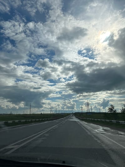 The scene depicts a wet asphalt road extending into the distance under a partially overcast sky in the vicinity of Petropavlovkskiy, Russia. Large cumulus clouds dominate the upper portion of the frame, with patches of blue sky visible and the sun brightly breaking through the clouds in the upper right quadrant. The road features double white lines in the center and shows significant wet areas, particularly on the right side, suggesting recent precipitation. To the left of the road, a flat, grassy expanse is visible, punctuated by numerous utility poles receding into the background, with distant structures or buildings faintly discernible on the horizon. To the right of the road, a metal guardrail with white and black stripes runs alongside. Beyond the guardrail, a grassy embankment leads to a treeline and two prominent lattice-structure power line towers. A sign with Cyrillic text is positioned between the two power towers. A faint reflection of a vehicle's dashboard is visible at the bottom edge of the image, indicating the photo was taken from inside a moving vehicle. A light-colored vehicle is observed in the far distance on the road. No individuals are explicitly visible.