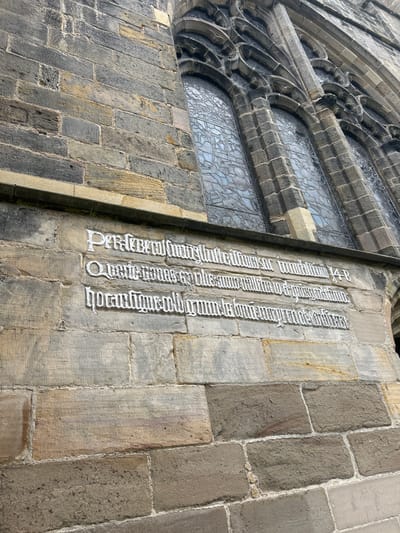 This image depicts a section of a building constructed from stone blocks, likely a church or similar historical structure, located in the city of Aberdeen City, United Kingdom. A window with intricate tracery is visible on the left side of the image. To the right, inscriptions are carved into the stone facade in a gothic style font. The first line appears to end with "multum 14-B", while the other lines are difficult to discern due to the font. The lighting suggests it is daytime, and the overall environment suggests an old and well-maintained building. There are no people visible in the image.