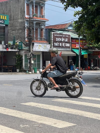 In Tân Quang Commune, Vietnam, a man wearing a light-colored helmet and a black t-shirt is riding a motorcycle with a gray seat and a black fender across a pedestrian crossing. He is wearing light blue shorts and flip-flops. The motorcycle has a silver exhaust pipe and a blue and white object attached to the rear fender. The background shows a street with buildings, trees, and utility poles. One building has a sign that says "COFFEE PIZZA" with an arrow pointing to the right. Another sign displays "SHOWROOM ĐỨC THỊN 2 GẠCH ỐP LÁT THIẾT BỊ VỆ SINH". The sky is overcast.