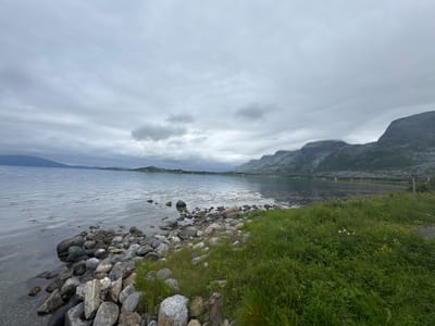 This image depicts a tranquil, natural landscape under an overcast sky. The foreground is dominated by a rocky shoreline bordering a calm body of water. The rocks vary in size and color, with some submerged in the shallow, clear water and others forming the bank. Lush green grass and some small wildflowers grow alongside the rocks, suggesting a temperate climate.
In the middle ground, the serene water stretches towards a distant, hazy shoreline. Mountains rise majestically in the background, their peaks partially obscured by clouds, creating a sense of depth and grandeur. The overall atmosphere is one of peaceful solitude.
The weather appears to be cool and cloudy, typical of a northern European climate. The diffused light suggests it is daytime, likely late morning or afternoon, with no direct sunlight. The scene evokes a feeling of calm and quiet contemplation. While no people are visible, the presence of a small signpost on the far right hints at a possible walking path or viewpoint, suggesting this is a place accessible to visitors. The location context of Sandnessjøen, Norway, aligns with the mountainous coastal scenery captured in the image. There is no visible text in the image.
