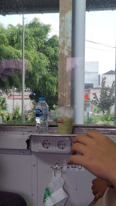 A person's hands are in the foreground, reaching towards electrical outlets. The background shows a window with trees and buildings visible outside, suggesting an urban or semi-urban environment. A bottle of water and a cup of green drink are placed on a shelf in front of the window. The outlets are mounted on a white panel. The overall scene appears to be inside a vehicle or a building with a window view. The mention of Karawang, Indonesia, is not directly supported by visual evidence in the image itself, but could be inferred from external context or metadata.