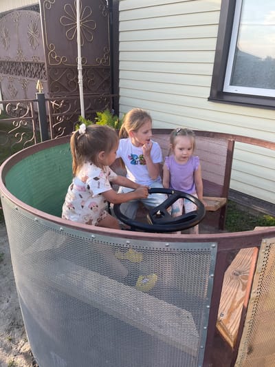 Three young girls are seated in a cylindrical, open-air play structure that resembles a ship's wheelhouse. The structure has a wooden frame and is covered with metal mesh on the exterior. A large black steering wheel is centrally positioned within the structure. The girl on the left, with her back to the camera, is wearing a white t-shirt with a cartoon print and is holding the steering wheel. She has a white ribbon in her hair. The girl in the middle, facing the camera and wearing a white t-shirt with a Stitch graphic, is looking towards the girl on the left. The girl on the right is wearing a purple t-shirt and patterned pants and is looking towards the front. The girls are sitting on a bench inside the structure. The background shows a beige-sided house with a window, a dark metal gate with decorative scrollwork, and greenery. The ground around the structure is covered with sand and some scattered vegetation. The scene appears to be outdoors in a residential area, likely a backyard, in the village of Mal'tsevo, Russia.