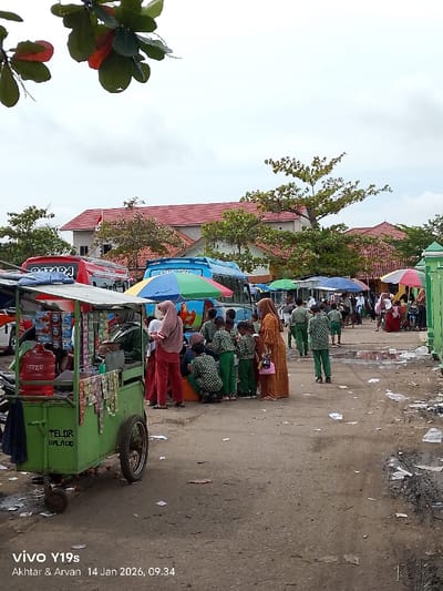 A street scene shows a group of children, mostly in green uniforms, gathered near a food cart. The cart, painted green, is selling "TELOR BALADO". Several adults are also present, some wearing hijabs. Buses are parked nearby, with colorful decorations. Umbrellas provide shade. A two-story building with a red tile roof stands in the background. The ground is uneven and appears to be dirt or gravel, with some scattered debris. The sky is overcast.