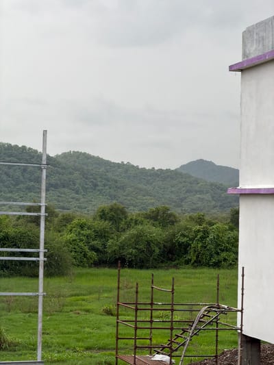 The image captures an overcast day scene in Vasai-Virar, India, featuring a lush green landscape juxtaposed with elements of construction. In the foreground, scaffolding made of rusted metal poles and horizontal bars is visible, with a wooden ladder section attached. To the right, a portion of a white building with a purple trim along its upper edge occupies the frame. A tall, slender metal pole stands to the left, with horizontal metal bars extending from it, suggesting it's part of a larger structure or utility pole.
Behind the immediate foreground, a wide expanse of vibrant green grass stretches out, dotted with scattered dry vegetation and small shrubs. Further back, a dense forest of trees with rich green foliage covers rolling hills, culminating in a backdrop of misty, layered mountains under a light gray, cloudy sky. The overall impression is one of a natural environment adjacent to ongoing construction work.