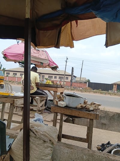 The image shows an outdoor market scene in Akute, Nigeria. A man in a yellow shirt sits at a wooden stall. The stall displays food items, including what appears to be boiled yam in a large pot. Bags and other objects are arranged around the stall. The stall is covered by a makeshift canopy of tarpaulins. A road and buildings are visible in the background. Overhead power lines are present.