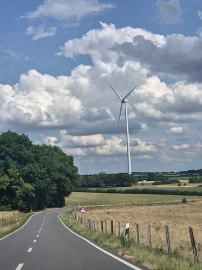 A tall, white wind turbine stands prominently in the background of a rural landscape. The turbine's blades are in a V-shape, suggesting they are stationary. The surrounding environment includes rolling fields of dry, golden grass, interspersed with patches of green vegetation and trees. A paved road curves from the foreground into the middle ground, with white dashed lines marking the center. A wooden fence with metal posts runs along the right side of the road. A triangular warning sign with a depiction of a skidding car is visible near the fence. The sky is filled with a mix of bright white and dark gray cumulus clouds, with patches of blue sky visible. The scene appears to be located in the municipality of Weiler-la-Tour, Luxembourg, given the context provided by the prompt. There are no people visible in the image.