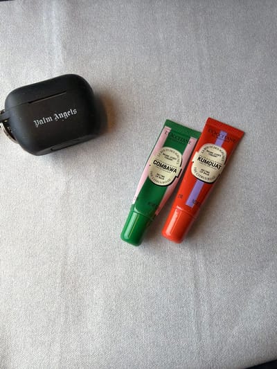 The scene is located in the city of Lykoshino, Russia. A black, rectangular object, identified as a "Palm Angels" branded case, is positioned in the upper left quadrant, featuring a metallic keyring on its left side. To the right, two L'OCCITANE EN PROVENCE "BAUME LÈVRES FONDANT" tubes are present. The left tube is green with a pink label section, identified as "COMBAWA" melting lip balm, containing 5% Shea butter and 12 ML volume. The right tube is red with a purple label section, identified as "KUMQUAT" melting lip balm, also containing 5% Shea butter and 12 ML volume. All objects rest on a light grey, textured surface, consistent with fabric. No individuals are present, and no actions or interactions are depicted beyond the static arrangement of these items.