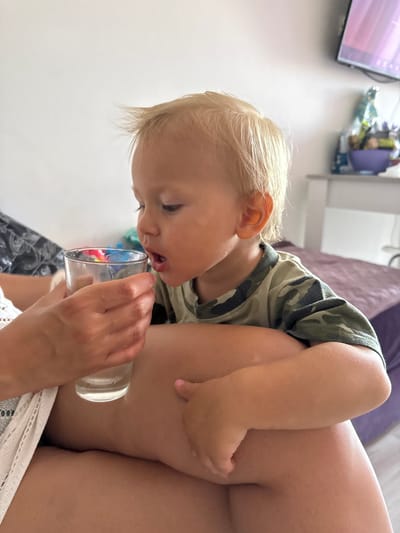 The image shows a close-up of a blonde toddler in a camouflage t-shirt being held by an adult. The toddler is leaning forward, attempting to drink from a clear glass that contains colorful small objects. The adult's hand is visible, holding the glass. The scene appears to be indoors, with a blurry background that includes what looks like a television screen and some shelves with items. The lighting suggests it might be daytime. The toddler's facial expression is focused on the task of drinking, and the overall mood seems casual and domestic. There is no visible text in the image. The location context is provided as Sochi, Russia.