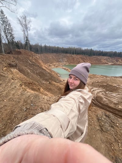 A woman stands on the edge of a large quarry in Ivlevo, Russia. She wears a tan jacket and a gray beanie. Behind her, the quarry walls are visible, leading down to a body of greenish-blue water. The sky is overcast.
