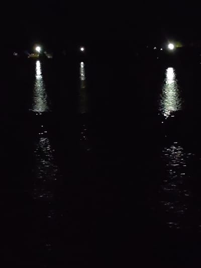 The image appears to depict a body of water, likely a lake or river, at night. There are three distinct light sources visible on the opposite shore, their reflections stretching across the water. The water's surface is disturbed, causing the light reflections to break up and scatter. The rest of the scene is dark, making it difficult to discern any further details about the surrounding environment. There is no information available about the location in the city of Zvoz, Russia.