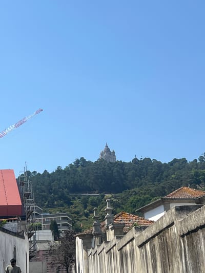 The image depicts a view of Viana do Castelo, Portugal, dominated by the Sanctuary of Santa Luzia, a prominent landmark situated atop a lush green hill. The sanctuary, with its distinctive dome and classical architecture, is visible in the distance, bathed in sunlight.
In the foreground, a weathered concrete wall runs across the frame, casting shadows and adding texture to the scene. To the left, a bright red roof of a building suggests ongoing construction or renovation, with scaffolding surrounding it. A tall construction crane, its boom extending towards the sky, is also present, indicating development in the area.
Further into the mid-ground, a multi-story building with a grey facade is nestled amongst the trees, contrasting with the natural landscape. The overall environment suggests a clear, sunny day with a bright blue sky overhead. The scene captures a blend of urban development and the natural beauty of the Portuguese coastline.