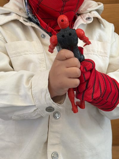 A child's hand grips a Spiderman action figure. The hand is placed on a white denim jacket. The action figure is red and black, with a web pattern. The jacket has silver buttons. The location is not Lermontovo, Armenia, as this information is not available in the image.