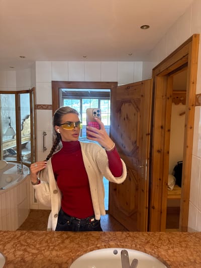 A mirror selfie is captured inside a bathroom or washroom in Going am Wilden Kaiser, Austria. A female subject is reflected from the chest to the upper thighs, wearing yellow-tinted sports sunglasses, white wireless earbuds, a red high-neck long-sleeved top, a cream knit cardigan with four large light-colored buttons, and dark denim jeans with a black belt. Her light-colored hair is styled in two braids, one of which she holds with her left hand, displaying multiple rings and a bracelet. Her right hand holds a light-colored smartphone with a pink textured case, pointed towards the mirror. The subject's gaze is directed slightly to her right. The environment features white tiled walls with a light brown decorative border, a mottled brown and orange countertop with a partially visible white porcelain sink, and recessed ceiling lights. To the left, a white corner bathtub or jacuzzi with chrome handles is visible. Two wooden doors are present in the background; the one on the right is partially open, revealing a room with wooden elements and a window overlooking a snowy, mountainous landscape characteristic of the Austrian Alps.