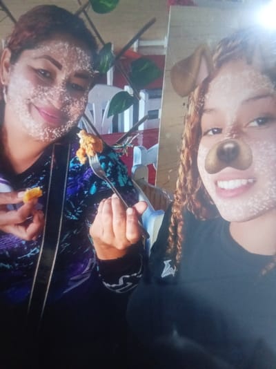 Two women are posing for a selfie. The woman on the left is holding a piece of food in one hand and a fork with food in the other. She is wearing a blue and purple top. The woman on the right has curly hair and is wearing a black shirt. Both women have a filter applied to their faces. There is a plant with green leaves in the background.