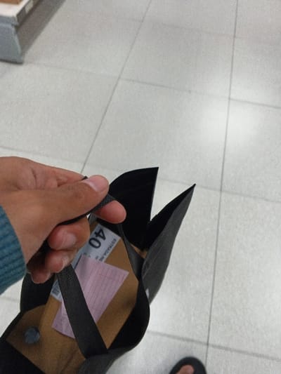A person holds a dark-colored shopping bag with a brown interior and a shipping label visible. The label displays the number "40" prominently. A pink sheet with grid lines is also inside the bag. The person's hand and a sleeve are partially visible. The background is a floor made of light-colored tiles. Part of a foot wearing a flip-flop is also seen in the lower right corner.