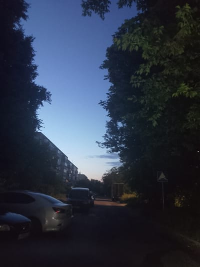 The image depicts a street scene at dusk in Lyubuchany, Russia. Two cars are parked on the right side of the road, one is white and the other is dark-colored. A light-colored truck is parked further down the road on the right. A pedestrian crossing sign is visible on the right side of the image. On the left side of the image, there is a multi-story apartment building. Trees are present on both sides of the road, with the foliage of the trees on the right side illuminated by what appears to be artificial light. The sky is a deep blue, indicating twilight.