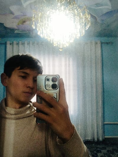 A young man takes a selfie indoors. He's wearing a beige turtleneck sweater and holding a phone with a metallic case. Behind him is a translucent white curtain. The room has a blue wall and ceiling with floral accents. A bright, ornate chandelier hangs from the ceiling, creating a strong light source in the room. The image appears to have a grainy quality.