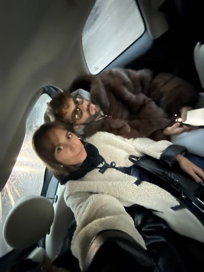 A man and a woman pose for a photo in a car. The man wears a brown fur coat and round sunglasses, holding a phone. The woman wears a white sherpa coat with black leather trim and a black scarf. A black purse sits beside her. Rain streaks the car window, and the interior is light gray. The image is taken at an angle, with the right side angled down.
