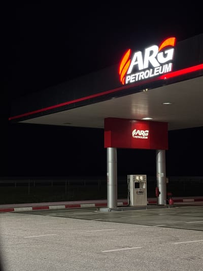 The image depicts an ARG Petroleum gas station. The station is illuminated with the brand's red and white signage. Gas pumps are visible along the roadside. The location is likely in or near Trilistnik, Bulgaria, given the brand is present there. The scene is observed at night with relatively clear weather. The area adjacent to the gas pumps appears to be paved. No individuals are visible in the scene.