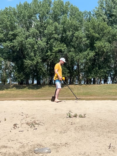 A man wearing a yellow t-shirt, white cap, and striped shorts is metal detecting on a sandy beach. He holds a shovel in his left hand and operates the metal detector with his right. Behind him, a line of lush green trees forms a backdrop against a clear blue sky. The sandy ground in the foreground is scattered with small plants and a discarded plastic bottle. The image appears to be taken on a sunny day. There is no visible indication of the location being in Indiya, Moldova.