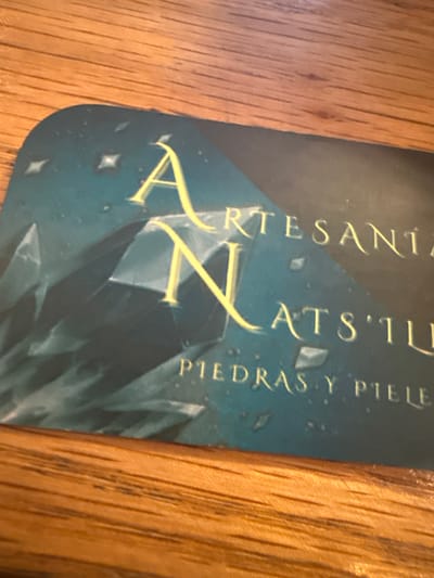 The image shows a business card for "Artesania Xats'il," subtitled "Piedras y Piele." It is photographed on a wood surface. There is no other information.