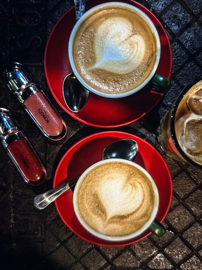 A top-down view captures a still life scene in Arthès, France, featuring an arrangement of beverages and cosmetic items on a dark, textured surface. Two white ceramic cups, each with a dark green handle, contain lattes with distinct heart-shaped latte art, resting on individual red saucers. A silver spoon is positioned on each saucer. To the left of the lattes, two KIKO Milano lip products are present; both are clear, cylindrical tubes with silver caps, one containing a shimmery peach-toned product and the other a shimmery reddish-brown product. On the right, a clear glass filled with iced coffee and ice cubes is partially visible. The underlying surface exhibits a dark, grid-like, possibly metallic or stone texture. No individuals are present in the frame.