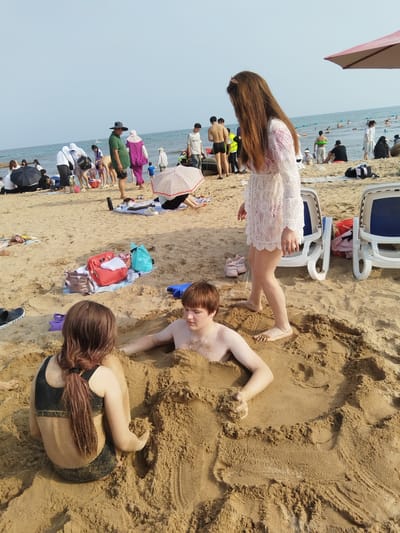 The image depicts a busy beach scene with numerous people enjoying a sunny day at Qinhuangdao, China. The main focus is on a group of individuals engaged in building sand sculptures and playing in the sand. Two young people, a boy and a girl, are partially buried in the sand, appearing to be having fun. The boy has his head and upper body exposed, while the girl's back is visible, with her head turned away from the camera. A woman in a white lace dress stands behind them, observing.
In the background, the beach is populated with many other beachgoers. Some are sitting or lying on the sand, while others are wading in the ocean. There are beach chairs, umbrellas, and various bags and personal belongings scattered on the sand. The sea is calm with a few people in the water, some with inflatable rings.
The time of day appears to be daytime, likely mid-morning to late afternoon, given the bright sunlight and clear sky. The weather is sunny and warm, typical for a beach day. The overall mood seems cheerful and relaxed, with people enjoying recreational activities.
Visible objects include beach chairs, umbrellas, bags (one red, one blue, and one beige), shoes (a black Crocs shoe is visible in the foreground), and inflatable rings in the water. A pink bow is in the hair of the woman in the white dress.
There is no visible text in the image, other than the brand name "Crocs" on the shoe. The location is identified as Qinhuangdao, China.