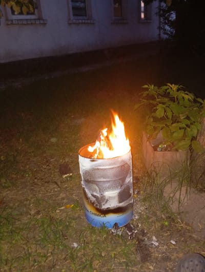 In Velyka Dymerka, Ukraine, an outdoor scene at night features a metal drum on fire. The drum is partially white and appears to be a discarded container. Bright orange flames erupt from the top of the drum, casting a warm glow on the surrounding area. The ground around the drum is covered in grass and some debris. In the background, a building with windows is visible, suggesting the scene is near a residential or industrial structure. A small bush with green leaves is to the right of the drum, and some small objects, possibly cans, are visible near its base. The overall atmosphere is dark, with the fire being the dominant light source.
