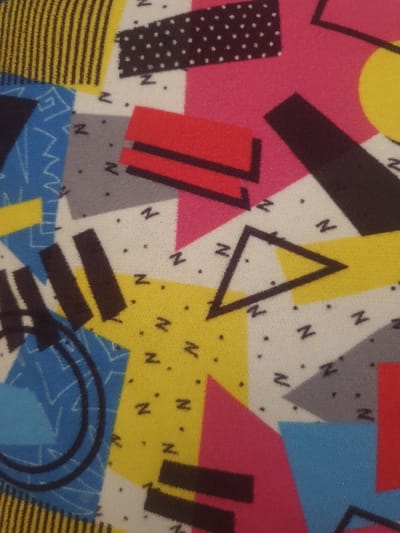 The image displays a textile surface featuring a multi-colored abstract geometric pattern. The design incorporates various shapes, including rectangles, triangles, and irregular polygons, rendered in a palette of white, black, yellow, hot pink, red, light blue, and grey. Specific elements include a black rectangle with white polka dots, areas of black vertical stripes on yellow, and red rectangular forms with black outlines. Scattered across white, yellow, and grey sections are small black dots and stylized 'Z' shapes. Other black linear elements present are a triangular outline and concentric circles. No individuals, animate objects, or environmental context beyond the pattern itself are depicted. This item is located in Mumbuni North ward, Kenya.