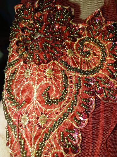 This image captures a close-up view of a richly embellished red textile, located in the city of Keo Kouang, Laos. The primary subject is a dense pattern of decorative elements stitched onto a red satin or silk-like fabric with a subtle sheen. The embellishments consist of numerous small, dark gold or bronze-colored spherical beads, forming intricate curvilinear designs and outlines. Interspersed among the beadwork are various iridescent, translucent red or fuchsia-colored sequins, generally circular in shape, which reflect light. Fine gold-colored metallic thread embroidery further defines patterns, including small, delicate star-like or web-like structures within the design. To the right of the ornate section, a plain, darker red mesh or net-like fabric is visible. No specific individuals, actions, or interactions are discernibly depicted within the frame. The environment is limited to the textile itself.