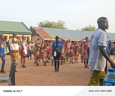 The image captures a scene in Gidan Kogi, Nigeria, featuring a group of children in traditional attire, likely participating in a cultural event. They are positioned in front of a person with their back to the camera, who is wearing a blue shirt with text. The children are all saluting with their right hands. A woman with a sash and crown is standing in the foreground. Buildings with corrugated metal roofs are visible in the background, with a tree line and overcast sky. A man in a gray shirt and yellow pants is walking to the right of the frame. There is a blue bottle in the lower right corner of the frame. There are people standing in the background behind the children and a white rope is used as a barrier between the children and the background. A black chair is present in the lower left of the image. The text at the bottom right reads "46mm f/1.8 1/407s ISO50", and "CAMON 40 Pro" is present in the lower left of the image.