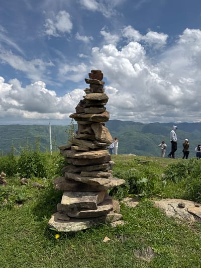 In Nizhnyaya Teberda, Russia, a stone cairn stands on a grassy hill overlooking a verdant mountain landscape under a partly cloudy sky. The cairn, constructed from numerous flat, irregular stones stacked vertically, is the primary subject in the foreground. Several people are visible in the mid-ground, standing on the elevated terrain, some appearing to observe the scenery. The environment features lush green grass, scattered wildflowers, and further back, rolling hills covered in trees, extending towards distant mountain ranges. A white fence runs horizontally across the middle ground, separating the foreground from the deeper landscape. The sky is a mix of blue and white, with fluffy clouds suggesting fair weather.