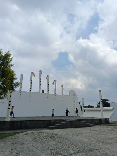 The image depicts an outdoor public space in Thành phố Biên Hòa, Vietnam, under an overcast sky. A large, predominantly white, multi-level concrete architectural structure occupies the middle ground. This structure features a tall, flat main wall adorned with at least eight stylized white cylindrical pillars, resembling flutes, complete with black circular markings (finger holes) and gold-colored cap-like elements at their tops. The structure includes an elevated concrete platform with steps leading up to it. Five young males are present on this platform. On the far left, one boy wearing a light top and blue shorts stands near a pillar. In the center, another boy in a dark shirt and light pants walks rightward. To his right, two boys, one in dark attire and another in a dark top and light shorts, are engaged near a small, light-colored ball, suggesting play. A fifth boy, dressed in a dark top and light shorts, stands further right on the platform. The foreground consists of a textured concrete ground surface. A portion of a green tree is visible on the far left, and distant vegetation appears behind the structure on the right. The sky is largely covered by white and light-gray cumulus clouds, with some patches of blue visible.