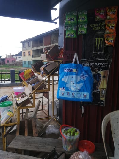The image depicts an outdoor vending stall in Ajasse Ipo, Nigeria. The stall features a display of various goods, including packaged snacks, beverages, and baby diapers. Prominently displayed is a large blue bag of "Kisskids Bai Diapers" and a can of "Monster Energy" drink. Other items visible include bags of "Viva Plus" detergent, packs of "Mama Joy," and a bucket filled with small, individually wrapped items, possibly candies or snacks. A yellow metal shelving unit holds several open cardboard boxes filled with products. In the background, a two-story building with balconies and balconies can be seen. The scene is outdoors, with a dark roof structure extending over the stall. A white plastic chair with the inscription "MRS" is positioned to the right of the frame. The ground appears to be paved or dirt, and a metal fence is visible in the background.