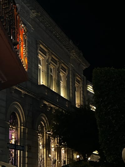An upward-angled nighttime view captures a multi-story, classically-styled building at the intersection of Plaza de la Paz and Calle Allende in Guanajuato City, Mexico. The building's upper facade displays at least five uniformly spaced rectangular windows, each emitting a warm, yellowish internal light. These windows are situated below a prominent, ornate cornice and above a continuous, shallow balcony supported by decorative corbels. The lower visible floor features a series of arched windows, two of which are clearly visible, adorned with intricate metal grates and illuminated internally with hues of purple and yellowish light. To the far left, a distinct orange-red light shines through a patterned screen, partially obscured by a dark, angled overhead structure. A white rectangular street sign, affixed low on the building, clearly reads "PLAZA DE LA PAZ" on its upper line and "ALLENDE" on its partially visible lower line. A dense, dark green mass of foliage, likely a large tree or hedge, occupies the right side of the frame, obscuring part of the building's right section. The sky is uniformly dark black, indicative of night. No individuals are present in the scene.