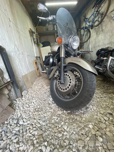 The image shows a cluttered garage or workshop. In the foreground, a large cruiser-style motorcycle with a windshield and spoked wheels is parked on a floor covered in gravel and small stones. The motorcycle is a light metallic color. To the right, the back end of another black motorcycle is partially visible. The walls of the structure appear to be made of concrete or brick, with some shelving and items stored on them. A bicycle is hanging from the ceiling on the right side of the frame. The lighting suggests it is daytime, with a fluorescent light fixture visible at the top. The overall impression is one of a working space used for storing vehicles and equipment. The location is identified as Znamensk, Russia.
