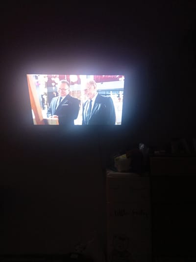 The image depicts a television screen displaying two men in suits, likely a still from a film or television program. The screen is illuminated, casting a glow onto the dark room. In the foreground, below the television, there is a dresser or cabinet with drawers. On top of the dresser, there are some soft objects, possibly stuffed animals or pillows, and to the right, a stacked set of drawers. The room appears to be dimly lit, suggesting it might be nighttime or the curtains are drawn. There is no discernible information in the image that would allow for identification of the location in Sibircevo, Russia.