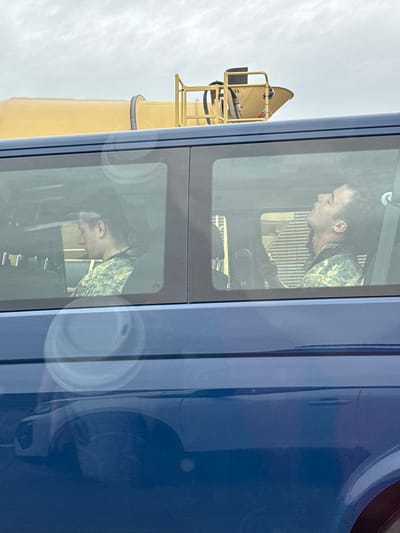 The image shows two men in military camouflage uniforms sitting inside a blue van. The man on the left is looking down and to the left. The man on the right is looking upwards, with his head tilted back. Both men appear to be in the passenger seats of the van. The van's windows reflect the sky and a yellow piece of machinery, possibly a construction vehicle or part of a building, in the background. The reflection also shows the front of another blue vehicle. The scene appears to be set in an outdoor urban or industrial environment. The location is the city of Bad Vöslau, Austria.