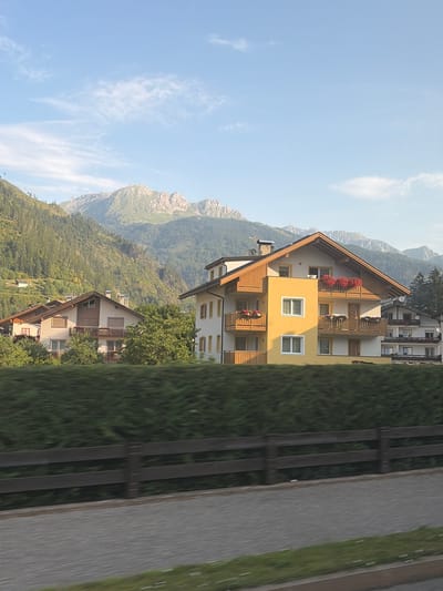 The image depicts a daytime scene in Predazzo, Italy, featuring the iconic Dolomites mountain range in the background. In the foreground, several buildings with a traditional Alpine architectural style are visible, characterized by steep roofs and wooden balconies. One prominent building is painted yellow and white, adorned with red flowers in window boxes. A dense green hedge runs horizontally across the middle ground, partially obscuring a wooden fence and a paved walkway. The sky is a clear blue with scattered white clouds. The overall impression is that of a serene mountain village during daylight hours.