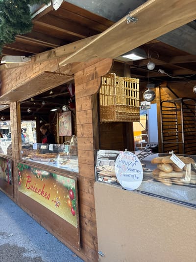 The scene depicts a wooden market stall, likely part of a Christmas market, located in Lucca, Italy. The stall is constructed from vertical dark-stained wooden planks, featuring an open service window with glass display cases. An awning made of light wood extends from the top.

Inside the stall, a single female vendor with light brown hair, wearing a black top and a red apron, is visible behind the counter, looking towards the left out of frame. The stall's interior ceiling has exposed wooden beams from which silver spherical Christmas ornaments are suspended. A red sign with "Joyeux Noel" written in white script hangs in the background. Wicker basket structures are mounted on the right interior wall.

The left display case contains numerous golden-brown baked goods, identifiable as "Brioches" by a decorative sign at the bottom of the stall, featuring a sparkly gold background with red Christmas ornaments and candy canes. The right display case holds multiple loaves of baguettes. A prominent white, starburst-shaped sign is affixed to this display case, stating "OFFERTA OGNI 3 BAGUETTES 1 GRATIS!" (Offer: Every 3 baguettes, 1 free!). A smaller white label next to the baguettes indicates "Mini Baguette 1€".

The immediate foreground shows a paved ground surface. In the upper left corner, green coniferous foliage is visible, consistent with seasonal decoration. The background, seen through the stall's opening, reveals bare trees and indistinct structures under a clear sky, indicating an outdoor setting.