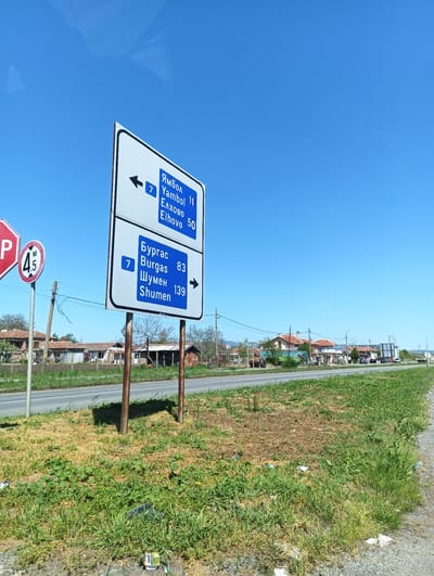 The image is a daytime shot of a road sign in what appears to be the rural outskirts of Zimnitsa, Bulgaria. The sign is white with blue text and arrows, indicating directions and distances to various cities: Yambol (11 km), Elhovo (50 km) to the left, and Burgas (83 km), Shumen (139 km) to the right. Both routes are on route 7. To the left of the road sign are two other signs, a red stop sign and a circular height restriction sign displaying "4.5 M". The road is flanked by grassy patches with scattered litter. Behind the road sign, a low row of modest houses is visible against a clear blue sky. Power lines run parallel to the road. The ground cover is a mix of dry and green vegetation.