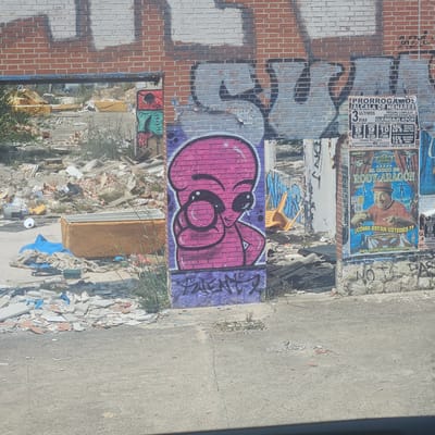 The image captures a dilapidated urban scene in Alcalá de Henares, Spain. A brick wall is covered in graffiti and faded posters, including advertisements for a local event, a KPOP event, and "El Circo de Rody Aragón". Debris and rubble are scattered on the ground, suggesting recent demolition or neglect. A yellow plastic chair sits amidst the wreckage. A concrete sidewalk extends to the right of the frame.