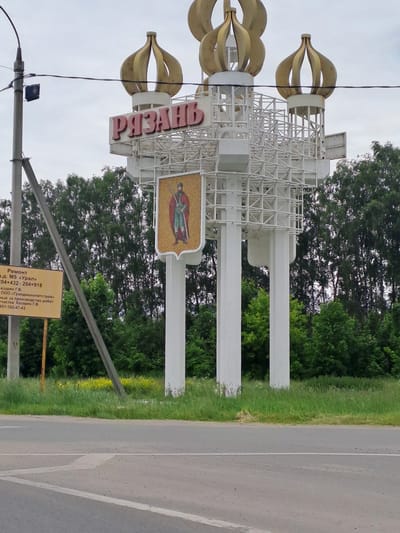 An image, originally rotated ninety degrees counter-clockwise, depicts a large, ornate roadside sign marking the entrance to the city of Ryazan, Russia, set against a backdrop of green trees and an overcast sky. The sign structure is predominantly white, featuring three large, golden, flame-like or stylized floral elements at the top. Below these, a white lattice framework supports the city's name, "РЯЗАНЬ" (Ryazan), rendered in prominent red capital letters. To the right of the name, a mosaic or painted emblem, shaped like a shield, displays a figure resembling a knight or warrior in a red cloak and green tunic, holding a weapon.
The sign is situated next to an asphalt road, visible on the right, which has a solid white line and a dashed white line marking lanes. Green grass and more trees are visible beyond the road and behind the sign structure. In the foreground, a utility pole with wires and a small black box is partially visible on the left.
Below the main sign, a smaller, rectangular yellow sign is present. Visible text on this sign indicates "Ремонт" (Repair/Maintenance) on "а.д. М5 «Урал»" (Federal highway M5 "Ural") between kilometer markers "204+432-204+918". It also mentions "Газарян Г.В." and "ООО «Грандмонолитстрой»" (LLC "Grandmonolitstroy") as being responsible for "производство работ" (work production) on a "частка Казарян Г.В." (plot/section Kazaryan G.V.), along with a phone number "951-102-47-43". The number "51" is also visible at the bottom right of this smaller sign. The scene appears to be taking place during daytime under an overcast sky.