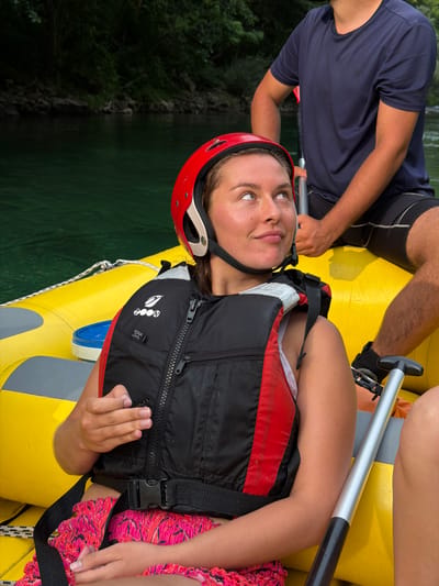 In Polje Bijela, Bosnia and Herzegovina, a young woman with brown hair is seated in a yellow raft on a body of water with a lush green forest backdrop. She is wearing a red helmet, a black and red life vest over a white top, and pink and black patterned shorts. Her left hand is raised, showing a butterfly resting on her index finger. To her right, another person, wearing a navy blue t-shirt and dark shorts, is also in the raft, holding a paddle. The water appears calm and the scene suggests a recreational activity on the river.
